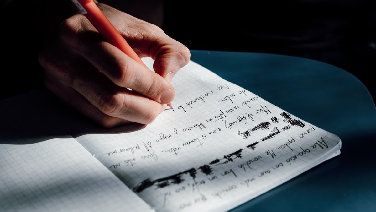 A person's hand writing in a notebook on a sunny day, showing detailed cursive notes.